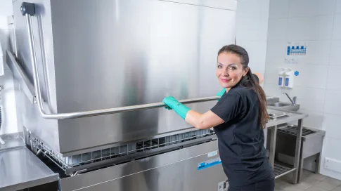 Frau in schwarzem T-Shirt arbeitet an einer großen Spülmaschine in einer Großküche.